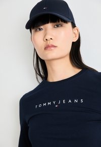 Fiatal nő tengerészkék sapkában és tengerészkék "Tommy Jeans" pólóban, előre néz, semleges arckifejezéssel, sima fehér háttér előtt.
