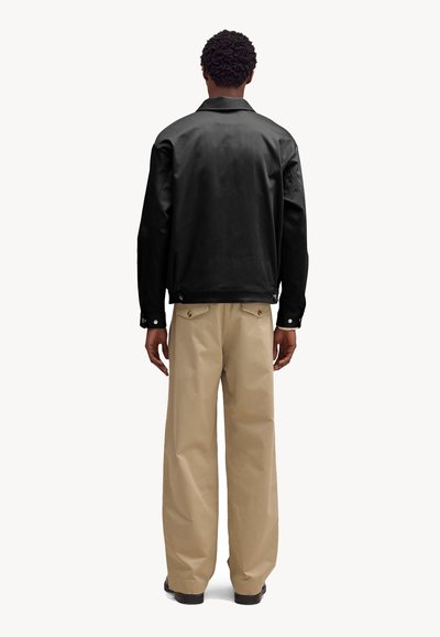 Uomo in piedi, visto di spalle, indossa una giacca nera e pantaloni beige a gamba larga, su uno sfondo bianco semplice.