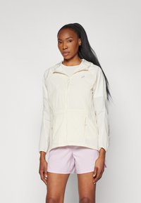 Leichte, cremefarbene Jacke mit Kapuze, Reißverschluss vorne, elastischem Bund und langen Ärmeln. Kombiniert mit rosa Shorts für einen lässigen Look.