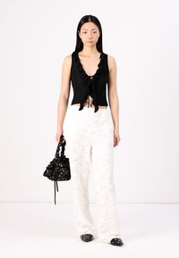 Top negru fără mâneci cu guler fronsat și detalii cu fundă, asortat cu pantaloni albi cu talie largă, având un model din dantelă florală. Geantă mică de mână neagră.