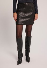 Czarna minispódniczka z cekinami o gładkiej fakturze, dopasowanym kroju i prostym dołem. Uzupełniona czarnymi kozaczkami na wysokim obcasie do kolan.