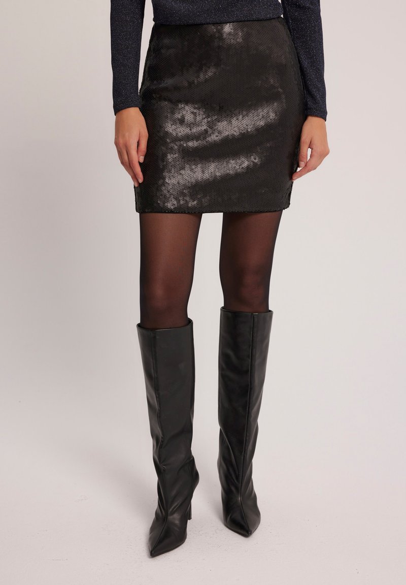 Czarna minispódniczka z cekinami o gładkiej fakturze, dopasowanym kroju i prostym dołem. Uzupełniona czarnymi kozaczkami na wysokim obcasie do kolan.