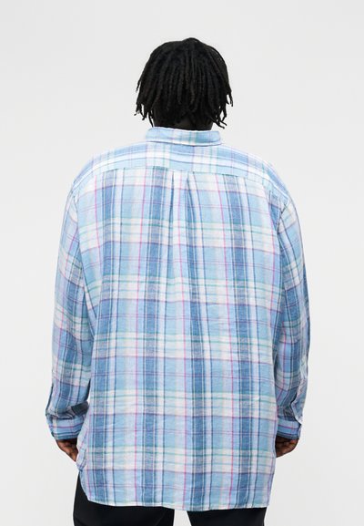 Persona con rastas cortas que lleva una camisa de manga larga, holgada, a cuadros azules y blancos, vista desde atrás.