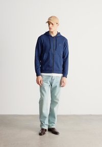 Tengerészkék cipzáras kapucnis pulóver húzózsinórokkal, fehér ingre húzva, világoskék farmerrel és barna cipővel párosítva. A megjelenést világosbarna sapka egészíti ki.