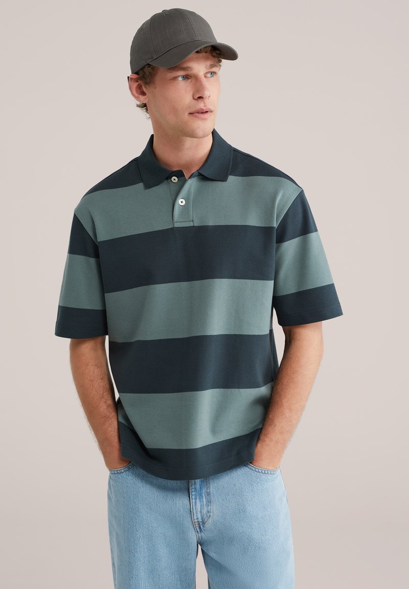 Jeune homme portant une casquette grise, un polo rayé vert et noir, et un jean bleu clair, debout les mains dans les poches, regardant vers la droite.