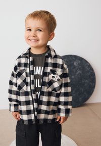 Schwarz-weiß karierte Jacke aus weichem Stoff mit einem Panda-Design auf der Tasche; darüber getragen ein dunkelgraues T-Shirt mit Grafikdruck.
