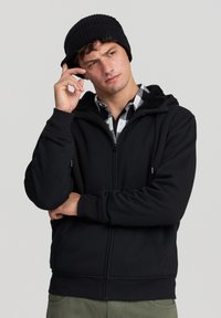 Felpa nera con zip e cappuccio foderato in pile, caratterizzata da zip frontale, tasche laterali e polsini a coste. Indossata sopra una camicia a quadri.