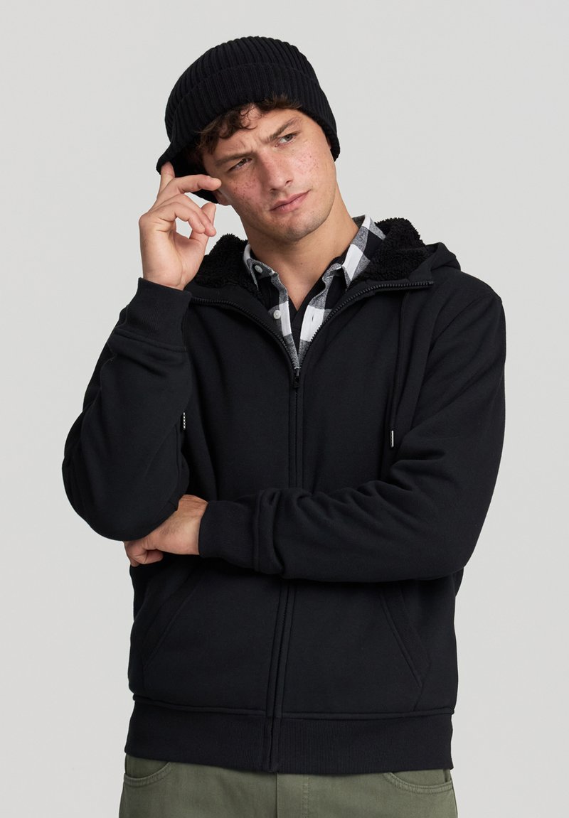 Felpa nera con zip e cappuccio foderato in pile, caratterizzata da zip frontale, tasche laterali e polsini a coste. Indossata sopra una camicia a quadri.