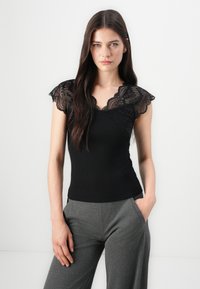 Top nero a coste con un design a maniche a cappuccio in pizzo, caratterizzato da un colletto scallop. Abbinato a pantaloni grigi a gamba larga.