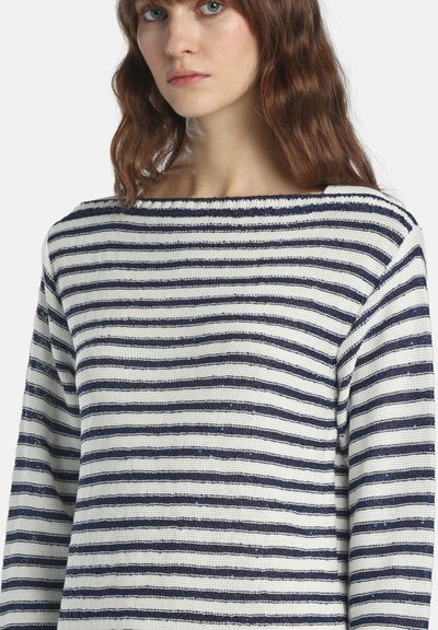 Femme aux cheveux bruns ondulés portant un pull blanc à rayures horizontales bleu marine avec des détails subtils scintillants.