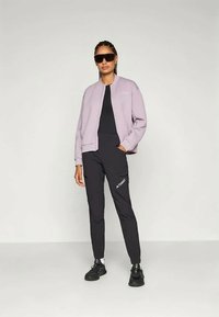 Helllila Bomberjacke aus strukturiertem Stoff, schwarzes Sportoberteil, schwarze Hose mit Logo und schwarze Sneakers, kombiniert mit Sonnenbrille.