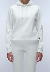 Persona che indossa un felpa bianca con cappuccio e pantaloni della tuta abbinati con loghi del marchio discreti sul petto e sulla coscia, in piedi su uno sfondo semplice.