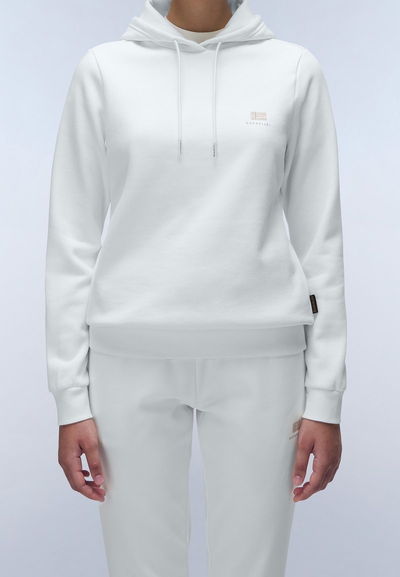 Persona che indossa un felpa bianca con cappuccio e pantaloni della tuta abbinati con loghi del marchio discreti sul petto e sulla coscia, in piedi su uno sfondo semplice.