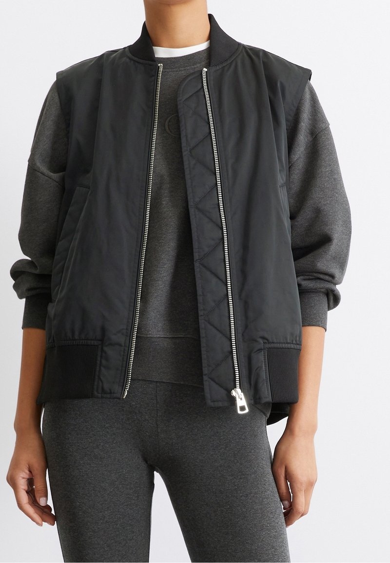 Zwarte bomberjack met doorgestikte details, ritssluiting aan de voorkant, geribbelde manchetten en een comfortabele pasvorm. Draagt over een grijze trui en bijpassende leggings.