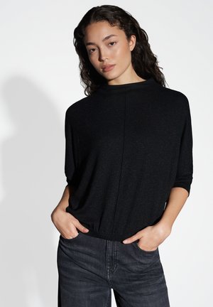 Zwarte, kortetermijn top met een hoge kraag en textuur met glinsteringen. Draag het met donkere jeans, handen in de zakken, wat een ontspannen pasvorm laat zien.