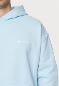 Hoodie azzurro chiaro realizzato in tessuto morbido; presenta una tasca a marsupio, polsini a costine e un logo bianco "PEGADOR" sul petto.