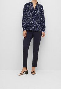 Marineblaue Bluse mit einem dezenten lila Blättermuster, mit V-Ausschnitt und langen Ärmeln. Kombiniert mit taillierten marineblauen Hosen und schwarzen Blockabsätzen.