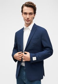 Νεαρός άνδρας φορώντας ένα ναυτικό μπλε σακάκι πάνω από ένα λευκό πουκάμισο και μπλε τζιν, κουμπώνοντας το σακάκι με τα δύο χέρια μπροστά σε ένα απλό φόντο.