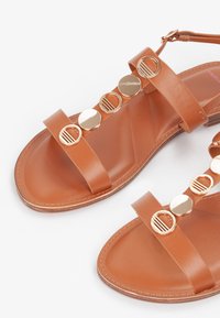 Sandalias de cuero marrón con suelas planas, que presentan acentos circulares dorados en las correas y una textura suave. Incluye una correa ajustable para el tobillo.