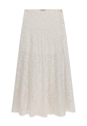 Frau mit langen, welligen Haaren, die einen weißen Spitzenrock und eine weiße Bluse trägt, steht neben einem rustikalen Holztisch in einem Raum im Vintage-Stil.