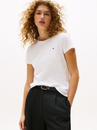 White cotton t-shirt with short sleeves, featuring a small logo on the left chest, paired with black tailored trousers and a gold-buckled belt.