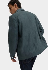 Uomo che indossa una giacca in suede verde scuro vista da dietro, abbinata a jeans blu scuro, su uno sfondo chiaro e semplice.
