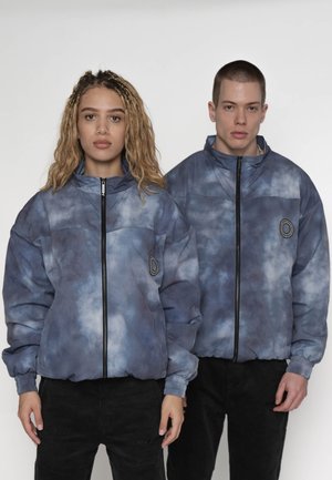 Dva modely, jedna žena a jeden muž, oblečení v zodpovedajúcich modro-šedých batikovaných bundách na zips a čiernych nohaviciach, stojaci vedľa seba.