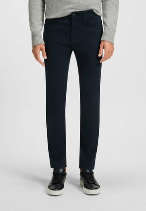 Mężczyzna w ciemnoniebieskich, dopasowanych spodniach, szarym swetrze i czarnych trampkach z białą podeszwą, stojący na jednolitym tle.