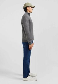 Pull gris zippé, jeans bleus et baskets blanches. Le modèle porte une casquette vert clair. Vue de profil avec un fond minimal.