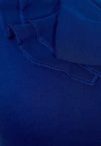 Blå tyg med ribbad textur och volangdetaljer vid halslinningen. Kanterna har en ren, avslutad söm.