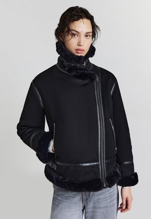 Giacca nera con colletto alto, caratterizzata da dettagli in pelliccia al colletto, ai polsini e all'orlo. Presenta cerniere argentate e un design leggermente oversize.