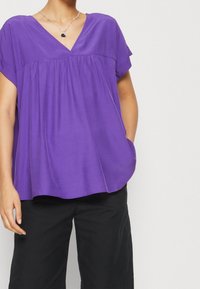 Lila kortärmad topp med V-ringning, med en samlad detalj under halsringningen och en lös, avslappnad passform. Matchas med svarta byxor.