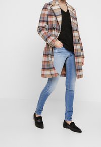 Beżowy, brązowy i granatowy płaszcz w kratę o podwójnym zapięciu, noszony na czarnej bluzie i niebieskich dżinsach, w zestawie z czarnymi loafersami.