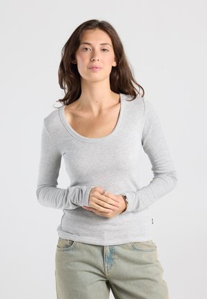 Jonge vrouw met lang bruin haar, een lichtgrijs shirt met lange mouwen en lichtblauwe spijkerbroek, staand met gevouwen handen.