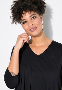 Schwarzes Kleid mit V-Ausschnitt und gefaltetem Design. Das Model hat lockige Haare und trägt auffällige Ohrringe. Glatte Stofftextur.