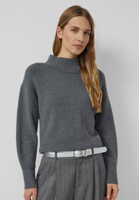 Wewnątrz szary sweter z dzianiny z wysokim ściągaczem, luźnym krojem i ściągaczami na rękawach, w połączeniu z srebrnym paskiem o fakturze i szarymi spodniami w paski.