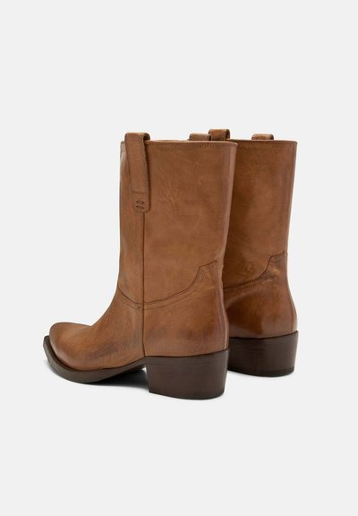 Bottes courtes en cuir marron avec une texture lisse, un bout arrondi et un petit talon bloc. Conçues avec un renfort à l'arrière et des languettes latérales pour faciliter l'enfilage.