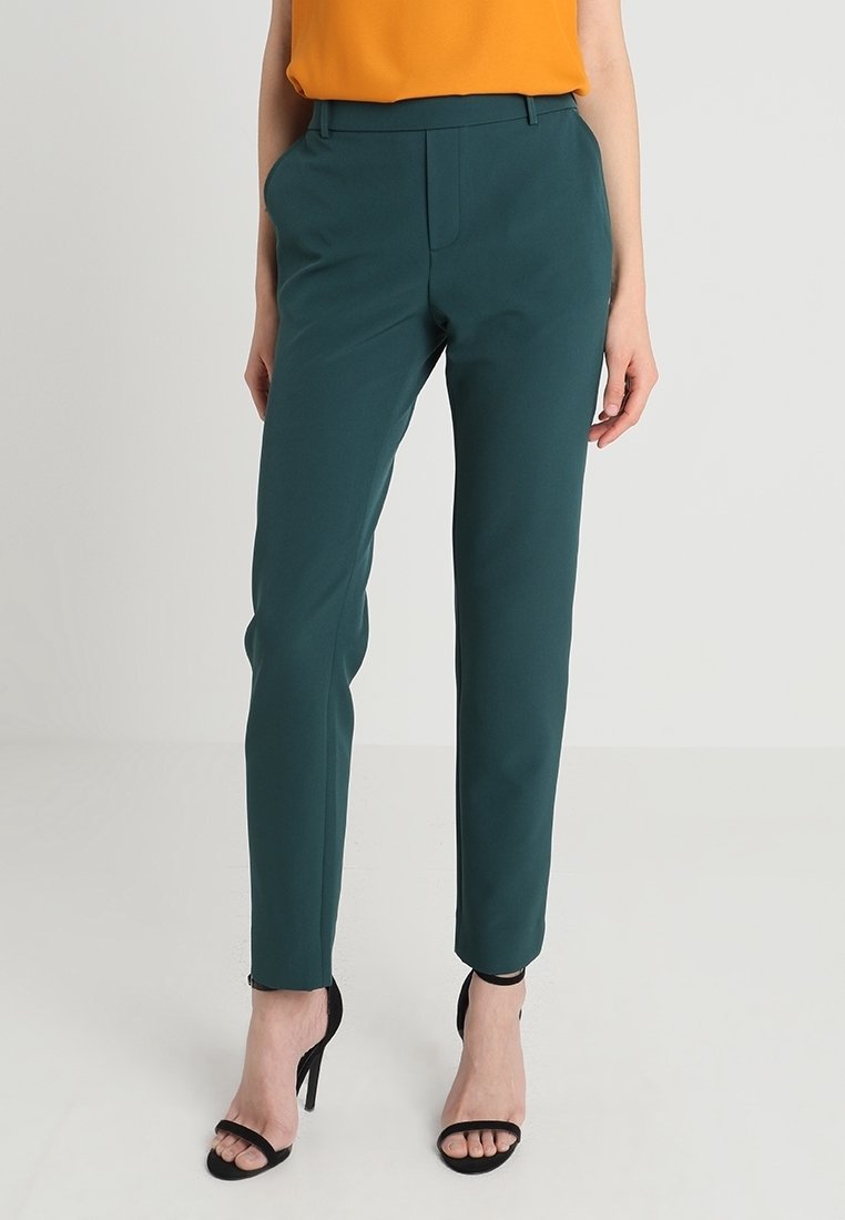 Groene op maat gemaakte broek met een smalle pasvorm, hoge taille, voorzakken en een gladde stoftextuur, gecombineerd met zwarte sandalen met bandjes en een oranje top.