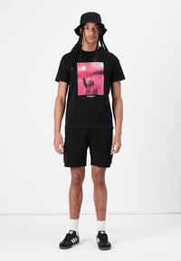 T-shirt noir avec imprimé graphique rose. Associé à un short noir. Les accessoires incluent un chapeau de bucket noir et des baskets noires.