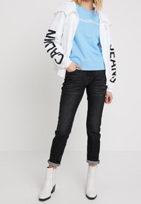 Bílá zipová bunda s černým nápisem, světle modrý svetr, tmavé denimové džíny a bílé kotníkové boty. Neformální outfit s vrstvenými texturami.