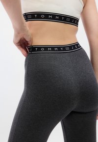 Γκρι κολάν με φαρδιά μαύρη μέση που φέρει το λογότυπο «TOMMY JEANS». Το υλικό φαίνεται μαλακό και ελαστικό με λεία υφή.