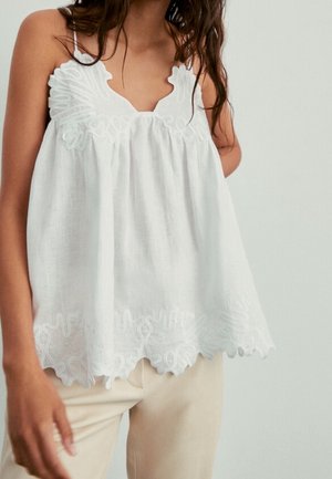 Woman wearing a loose white sleeveless top with floral embroidery and scalloped edges, paired with light beige pants.