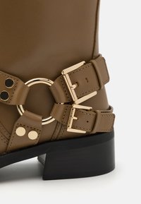 Bocanci din piele maro, până la gleznă, cu două catarame aurii, un accent metalic circular și un toc mic, negru, de tip block. Textură netedă cu detalii de cusătură.