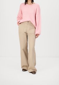 Un maglione rosa lavorato a maglia con maniche oversize abbinato a pantaloni beige a vita alta e a gamba larga, caratterizzati da cuciture minime e una vestibilità rilassata.