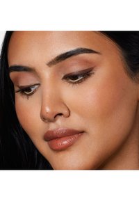 Gros plan sur un visage avec des yeux marron, une peau bronzée et des lèvres douces et brillantes. Le maquillage des yeux présente un fard à paupières aux nuances chaudes et des sourcils bien dessinés.