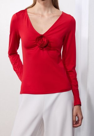 Femme portant un haut rouge à manches longues avec un col en V et un détail de rose en tissu au centre de la poitrine, ainsi qu'un pantalon blanc sur un fond uni.
