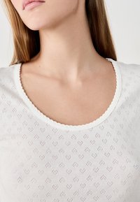Witte top met een ronde halslijn, gekenmerkt door een textuurpatroon van kleine hartjes en een golvende rand rond de kraag.