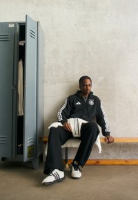 Czarny hoodie Adidas z trzema białymi paskami, czarne spodnie i białe buty z czarnymi akcentami. Siedzi na drewnianej ławce obok szarej szafki.