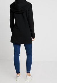 Zwarte gestructureerde jas met een grote capuchon, gecombineerd met blauwe skinny jeans en witte sneakers. De jas heeft een getailleerd ontwerp en lange mouwen.