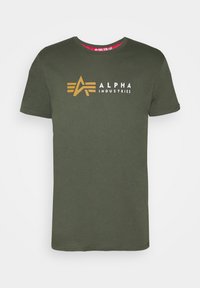 Grønn t-skjorte med kort erme og rund hals, med gul Alpha Industries-logo og hvit tekst midt på brystet.
